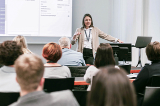 Referentin Elisabeth Freitag steht vor einem Bildschirm und unterrichtet mehrere Personen. 